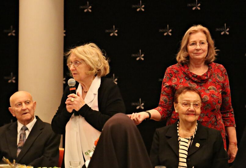 Zdjęcie: kobieta z mikrofonem i grupa osób siedzących przy stole w odświętnych strojach