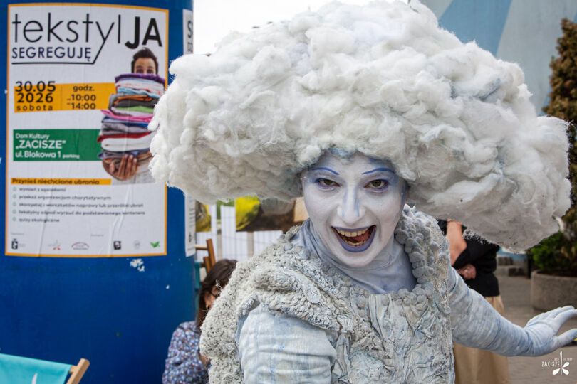 Zdjęcie: osoba w teatralnym kostiumie przypominającym chmurę, z białym makijażem i dużą puszystą peruką, za nią plakat na słupie z napisem Tekstylia segreguję