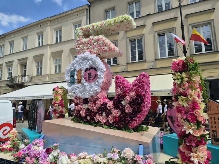 Zdjęcie: instalacja artystyczna z kwiatów, w jasnych kolorach, symbolizująca warszawską syrenkę, w tle kamienice